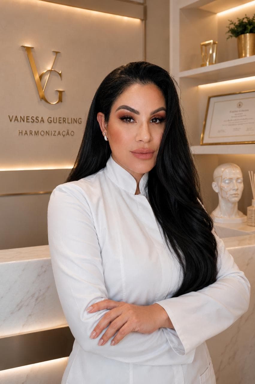 Vanessa Guerling de jaleco branco no consultório, ao lado da parede com a marca Vanessa Guerling Harmonização e o certificado de registro emoldurado.